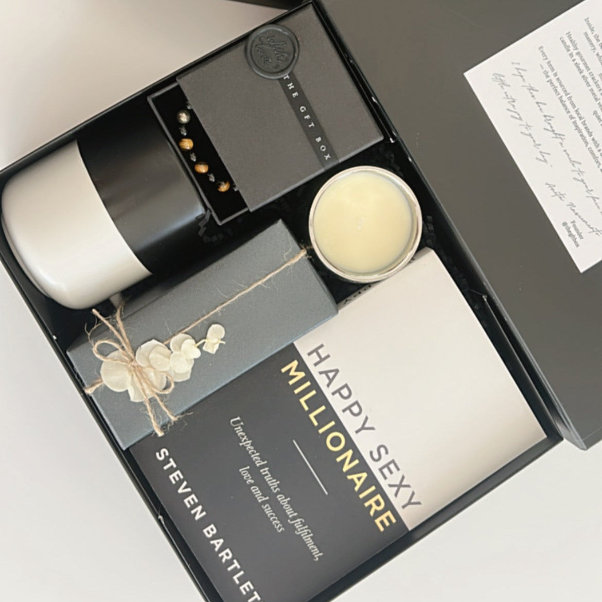 Ready to go gift set with a book titled 'Happy Sexy Millionaire' by Steven Bartlett, a candle, a mug, a crystal bracelet for men and a box of crackers. Packaged and wrapped readymade gift for men.