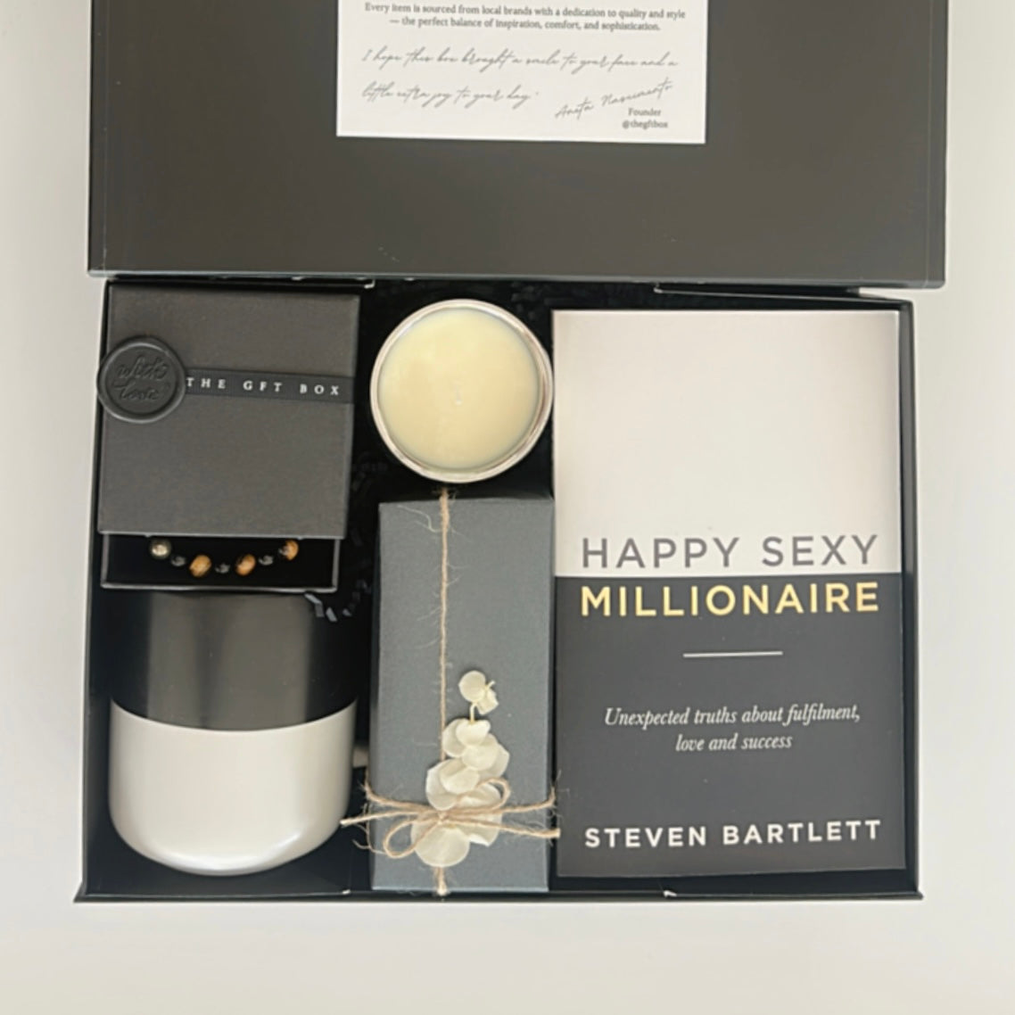 Ready to go gift set with a book titled 'Happy Sexy Millionaire' by Steven Bartlett, a candle, a mug, a crystal bracelet for men and a box of crackers. Packaged and wrapped readymade gift for men.