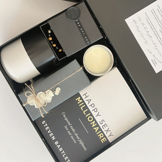 Ready to go gift set with a book titled 'Happy Sexy Millionaire' by Steven Bartlett, a candle, a mug, a crystal bracelet for men and a box of crackers. Packaged and wrapped readymade gift for men.